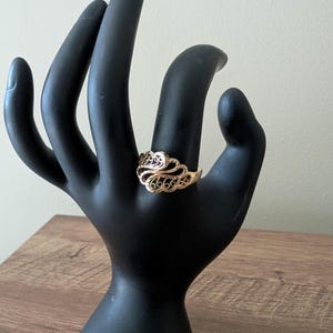 May include: A gold-colored ring with an ornate, filigree design. The ring features a series of swirling patterns and small, repeating shapes. The ring is displayed on a black hand-shaped display stand, set against a neutral background.