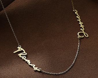 May include: A gold-coloured necklace with the names "Thomas" and "Jessica" in a cursive script. The names are linked by a fine chain, creating a personalised jewellery item. The necklace is displayed on a brown surface with a wooden accent.