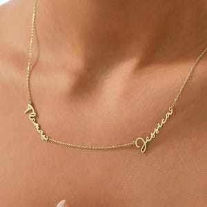 May include: A delicate gold-colored necklace featuring two names in a cursive font: "Teresa" and "Jessica." The necklace is worn around the neck, with the names positioned on the chest. The chain is thin and elegant, suitable for everyday wear.