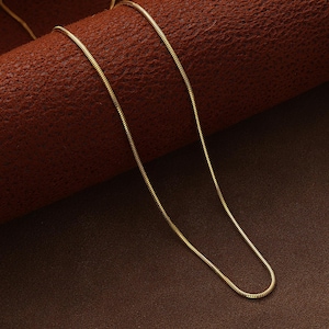 May include: A fine gold-coloured snake chain necklace. The chain is slender and flexible, with a smooth, reflective surface. The necklace is displayed on a brown surface, highlighting its warm, metallic tone. A simple and elegant piece of jewellery.