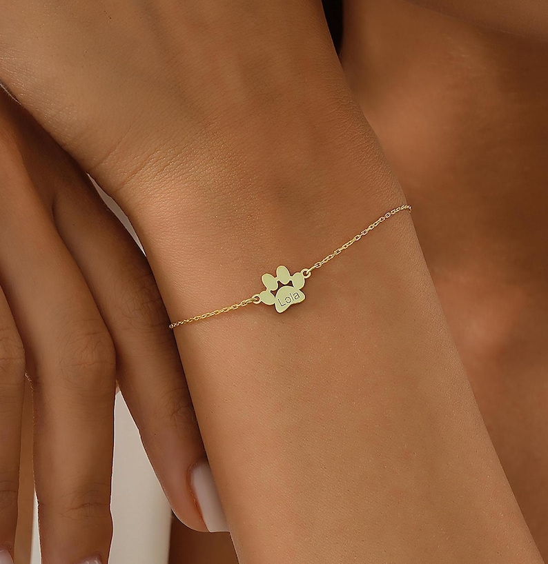 May include: A gold chain bracelet with a paw print charm engraved with the name "Lola".