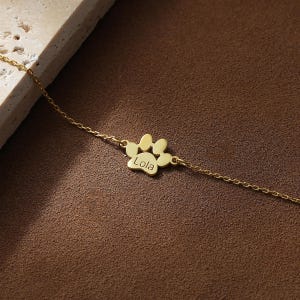 May include: A gold-colored bracelet featuring a paw print charm with the name "Lola" engraved on it. The delicate chain and charm are displayed against a brown background, highlighting the jewelry's design.