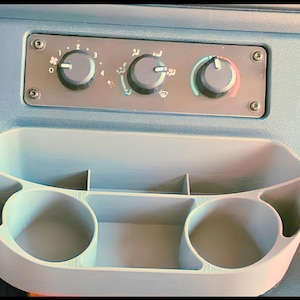May include: A light blue car dashboard with air conditioning controls and a storage compartment. The dashboard features three circular dials for fan speed and air direction, along with a built-in cup holder and storage spaces.