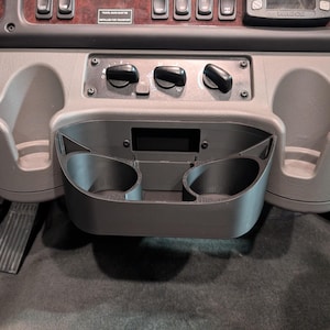 May include: A gray plastic cup holder with two cup slots. The cup holder is mounted on a gray dashboard with a black control panel.