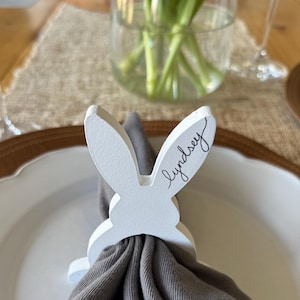 May include: A white wooden bunny-shaped napkin ring with the name "Lyndsey" written on it, holding a gray napkin. The napkin ring is on a white plate, with a glass of water and a vase of flowers in the background.