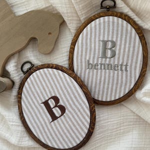 May include: Two oval embroidery hoops with wooden frames. Each hoop features a striped fabric background and embroidered text. One says "B" and the other says "bennett". The hoops are displayed on a white, textured surface.
