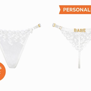 Op de afbeelding: Witte kanten string met verstelbare bandjes en goudkleurige hardware. Eén string heeft het woord "BABE" in goudkleurige letters. Het woord "GEPERSONALISEERD" staat in een oranje banner.