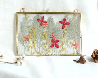 May include: A rectangular, gold-framed pressed flower art piece. The artwork features red, yellow, and white flowers and greenery. A gold chain is attached to the frame. A small brown sculpture and a pinecone are next to the frame.
