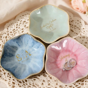 May include: Three flower-shaped ceramic trinket dishes in light blue, pale pink, and mint green. Each dish features gold-colored custom text and dates. The pink dish holds two silver-toned metal rings. The dishes have a glossy, glazed finish.