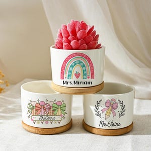 May include: Three white ceramic planters with bamboo saucers. One planter features a rainbow design with the text "Mrs. Miriam", another has a gift box design with "Mrs. Connie", and the third has a pencil and bow design with "Mrs. Elaine". A red succulent sits in the top planter.