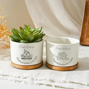 May include: Two white ceramic planters with bamboo saucers. One planter holds a green succulent. Both planters have the text "First Home" and a house graphic. One reads "Thomas & Stella 12.23.2025" and the other reads "Angel & Kinsley 05.13.2025".