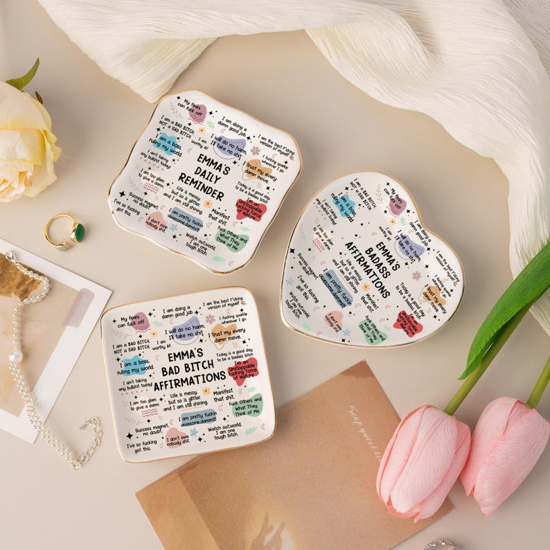 May include: Three ceramic trinket dishes with gold rims. One is square, one is heart-shaped, and one is rectangular. Each dish has a different design with the text "EMMA'S DAILY REMINDER" or "EMMA'S BAD BITCH AFFIRMATIONS".