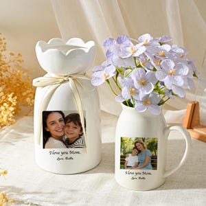 May include: Two white ceramic vases, one with a scalloped top and ribbon, the other a pitcher with a handle, each displaying a photo and the text "I love you Mom." The pitcher holds light purple flowers. A clear sign reads "The best yet to come."