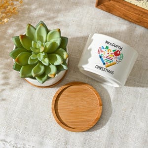May include: A white ceramic planter with a green succulent plant, a wooden saucer, and a second white planter with the text "Mrs. Darcy CHRISTMAS" and a heart-shaped design of school supplies. The succulent is in a white pot.