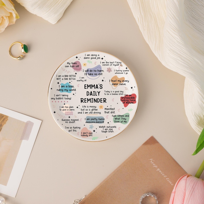 May include: A white ceramic daily reminder with the text "EMMA'S DAILY REMINDER" and various motivational phrases. The round plate features a gold rim and is surrounded by other decorative items.