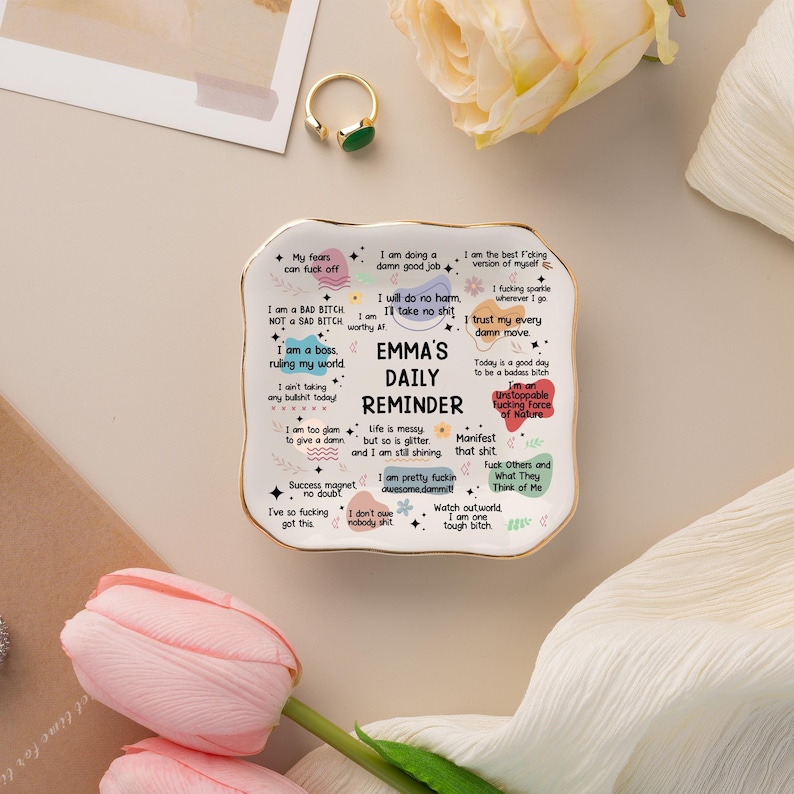 May include: A white ceramic daily reminder dish with a gold rim, featuring the text "Emma's Daily Reminder" and various motivational phrases. The dish is rectangular with rounded edges and is surrounded by flowers and a ring.
