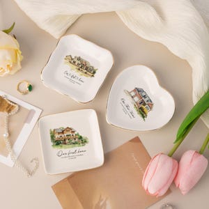 May include: Three white ceramic trinket dishes with gold-trimmed edges. Two dishes are square and one is heart-shaped. Each features a watercolor illustration of a house and the text "Our first home" with specific location and date details.