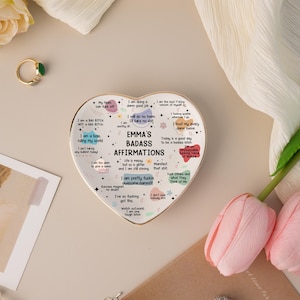 May include: A heart-shaped ceramic dish with a gold rim, featuring the text "EMMA'S BADASS AFFIRMATIONS" and various positive affirmations. The dish is white with colorful text and graphics. A gold ring with a green stone is visible.