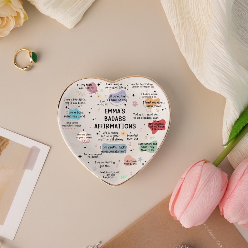 May include: A heart-shaped ceramic dish with a gold rim, featuring the text "EMMA'S BADASS AFFIRMATIONS" and various positive affirmations. The dish is white with colorful text and graphics. A gold ring with a green stone is visible.
