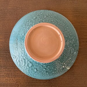 May include: A ceramic bowl with a textured, turquoise exterior and a light brown interior. The bowl is round and appears to be handmade. The underside of the bowl is visible, showing the unglazed base.