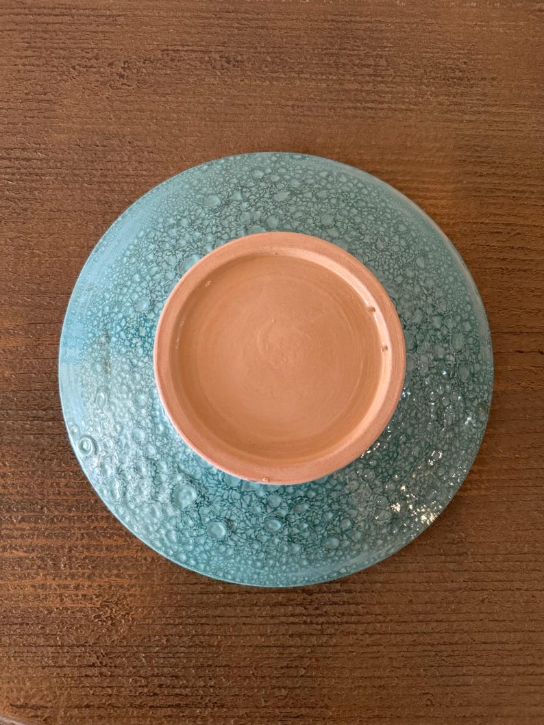 May include: A ceramic bowl with a textured, turquoise exterior and a light brown interior. The bowl is round and appears to be handmade. The underside of the bowl is visible, showing the unglazed base.