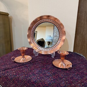 May include: A copper-toned vanity set featuring a round mirror with a decorative border and two matching candle holders. The mirror is propped up on a clear stand, and the candle holders have a handle. The set is displayed on a patterned cloth.