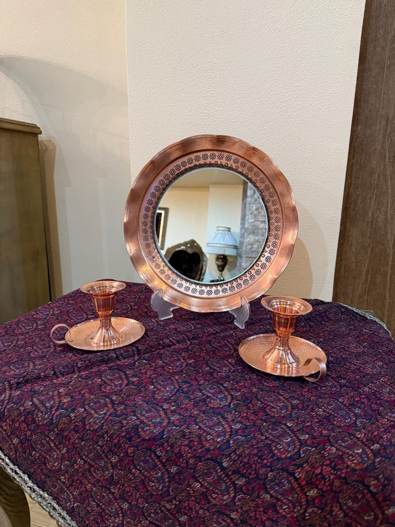 May include: A copper-toned vanity set featuring a round mirror with a decorative border and two matching candle holders. The mirror is propped up on a clear stand, and the candle holders have a handle. The set is displayed on a patterned cloth.
