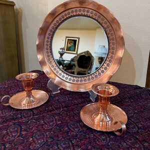 May include: A copper-colored decorative mirror with a floral patterned border, and two matching candle holders. The mirror reflects a room with furniture and artwork. The candle holders have a handle and a textured base.