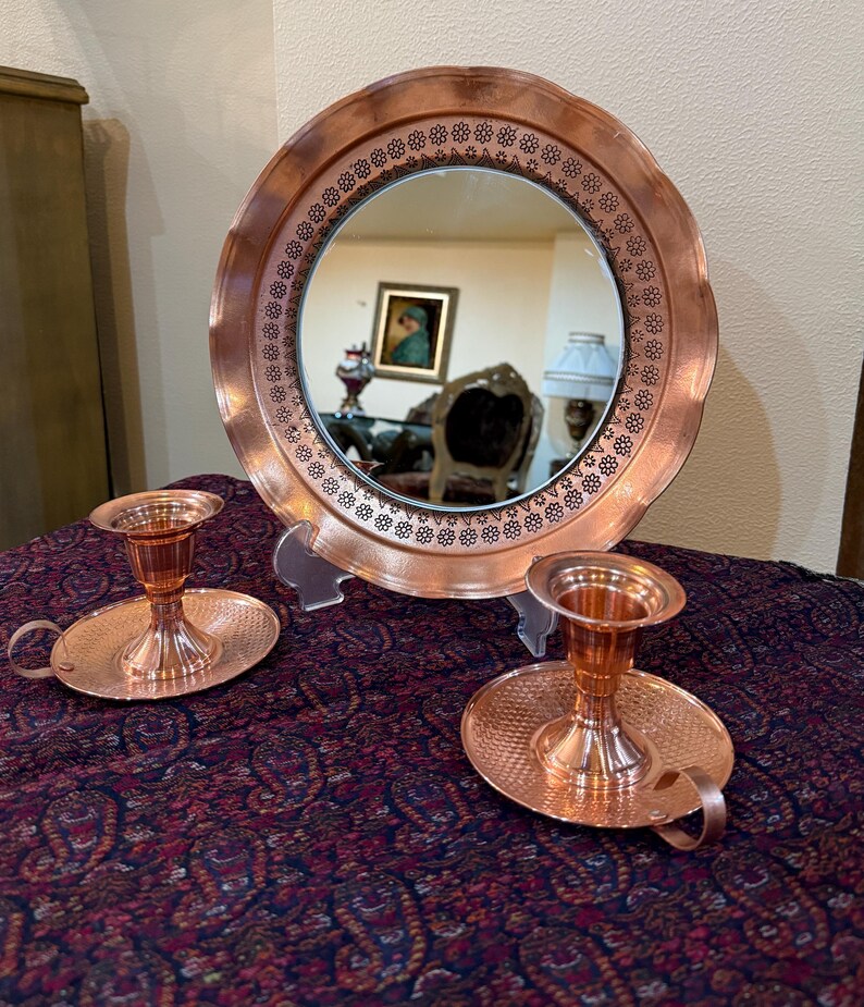 May include: A copper-colored decorative mirror with a floral patterned border, and two matching candle holders. The mirror reflects a room with furniture and artwork. The candle holders have a handle and a textured base.