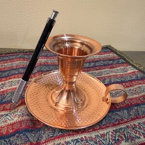 May include: A copper candle holder with a round base, a central cup, and a handle. The base has a textured pattern. A black and silver mechanical pencil rests on the side. The candle holder is on a patterned textile.