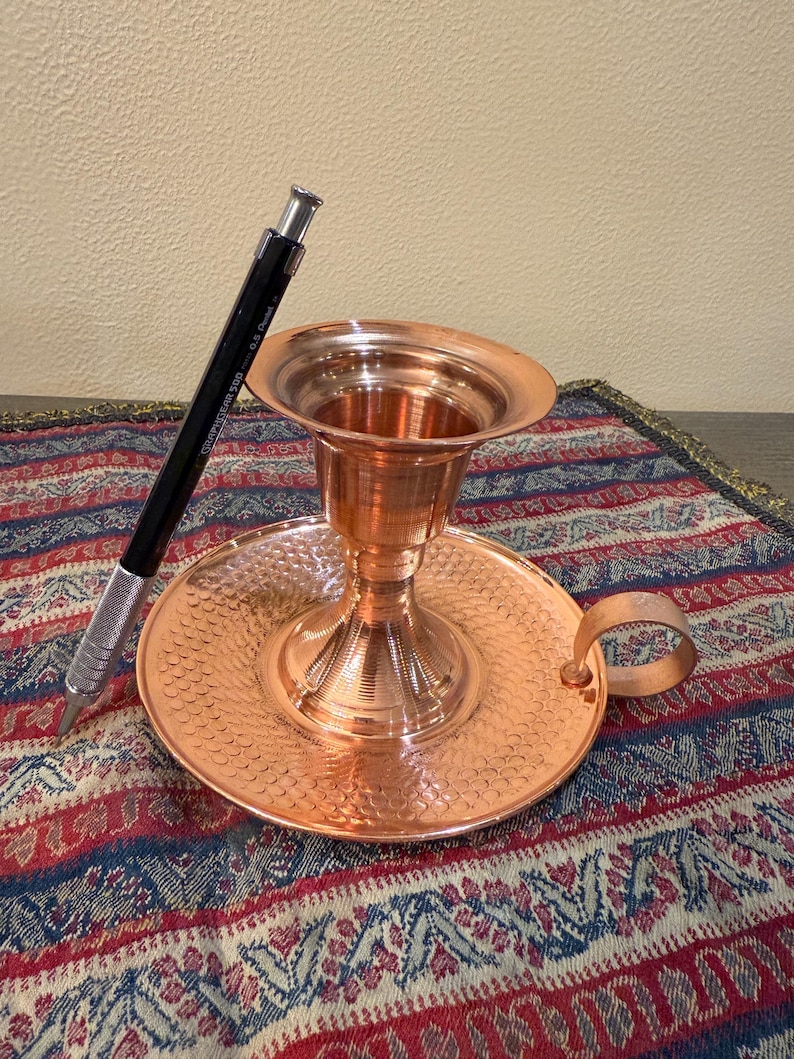 May include: A copper candle holder with a round base, a central cup, and a handle. The base has a textured pattern. A black and silver mechanical pencil rests on the side. The candle holder is on a patterned textile.