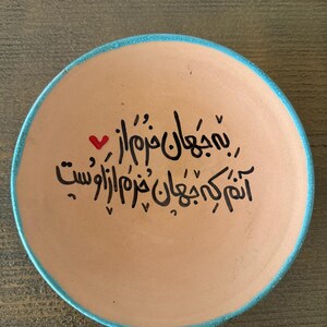 May include: A round, tan ceramic plate with a turquoise blue rim. The plate features black calligraphy and a small red heart. The text is in a script style and covers most of the plate's surface.