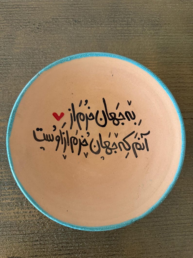 May include: A round, tan ceramic plate with a turquoise blue rim. The plate features black calligraphy and a small red heart. The text is in a script style and covers most of the plate's surface.