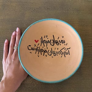 May include: A round, terracotta-colored plate with a turquoise rim and black calligraphy. A small red heart is next to the calligraphy. The plate is held by a hand with a silver ring.