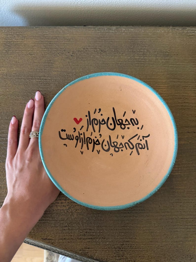 May include: A round, terracotta-colored plate with a turquoise rim and black calligraphy. A small red heart is next to the calligraphy. The plate is held by a hand with a silver ring.