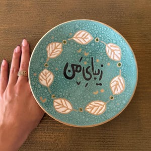 May include: A round, turquoise ceramic plate with a leaf design and text in black. The plate has a white leaf pattern around the edge and is held by a hand.