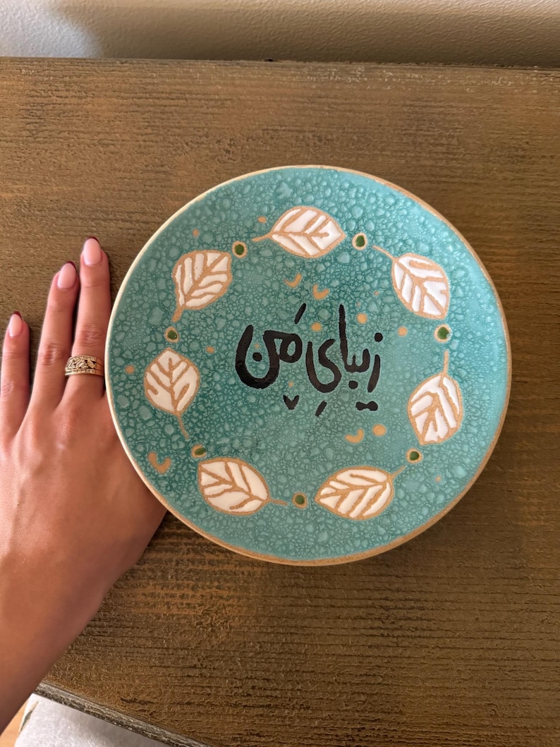 May include: A round, turquoise ceramic plate with a leaf design and text in black. The plate has a white leaf pattern around the edge and is held by a hand.
