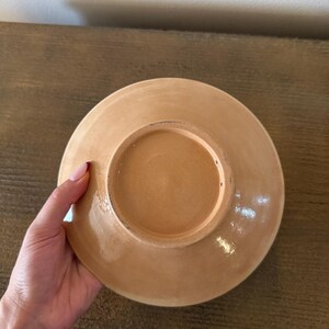May include: A ceramic plate with a light brown glaze. The plate is round with a slightly raised edge and a recessed center. The underside of the plate is visible, showing the unglazed base and a circular indentation.