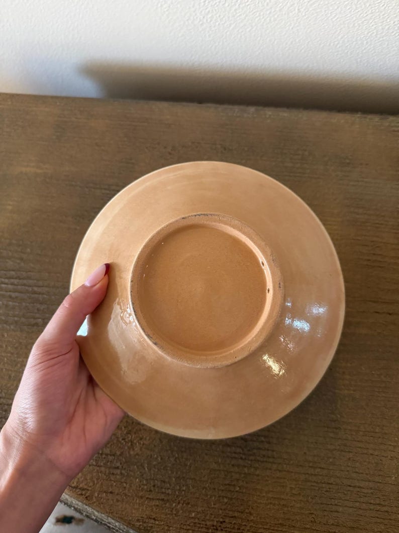 May include: A ceramic plate with a light brown glaze. The plate is round with a slightly raised edge and a recessed center. The underside of the plate is visible, showing the unglazed base and a circular indentation.
