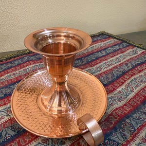 May include: A copper candlestick holder with a wide, flared top and a circular base. The base has a textured, hammered design. A small handle is attached to the side. The candlestick holder is displayed on a patterned textile.