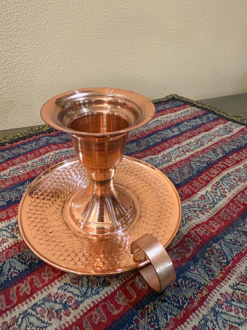 May include: A copper candlestick holder with a wide, flared top and a circular base. The base has a textured, hammered design. A small handle is attached to the side. The candlestick holder is displayed on a patterned textile.