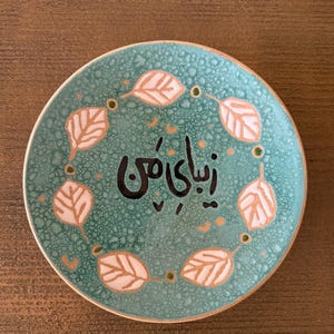 May include: A round, turquoise ceramic plate with a textured surface. The plate features a ring of white and gold leaf designs around the edge, with the black Persian text "Zibaye Man" in the center. The plate is on a brown wooden surface.