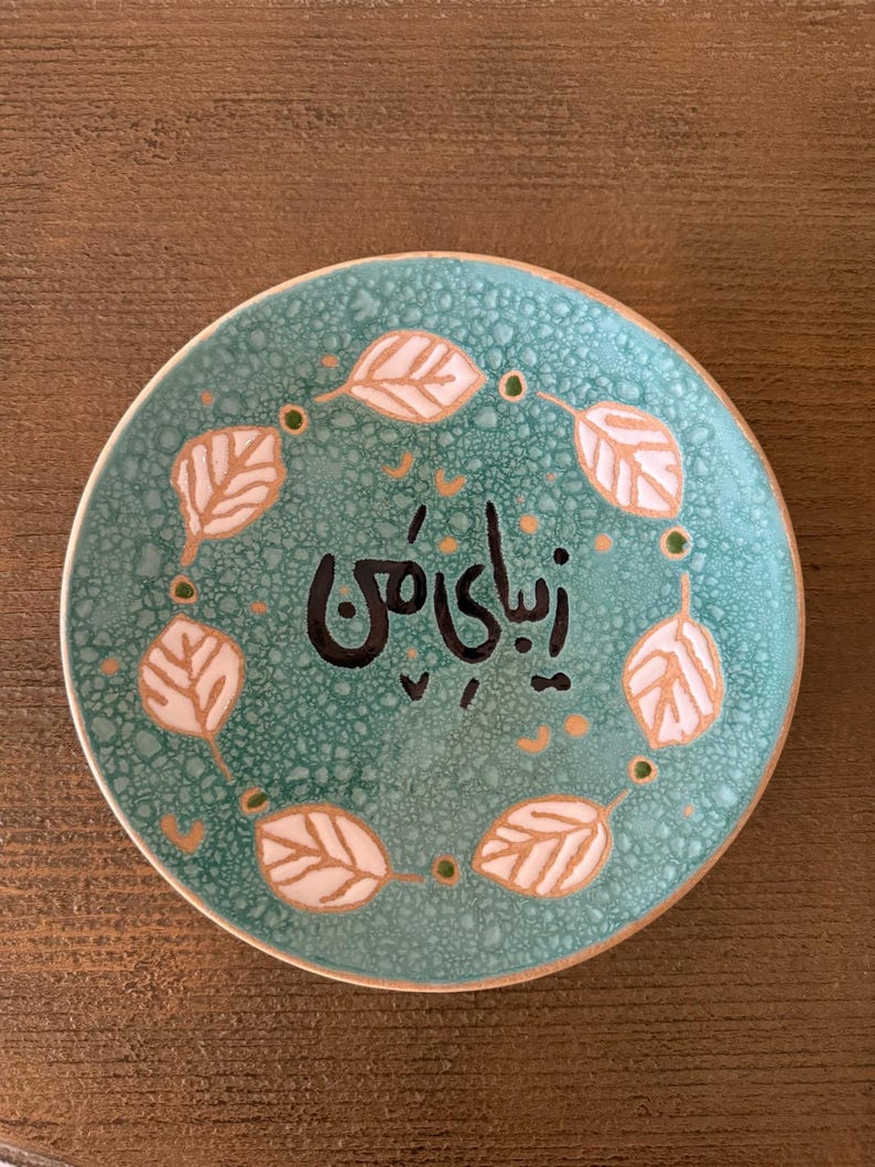 May include: A round, turquoise ceramic plate with a textured surface. The plate features a ring of white and gold leaf designs around the edge, with the black Persian text "Zibaye Man" in the center. The plate is on a brown wooden surface.