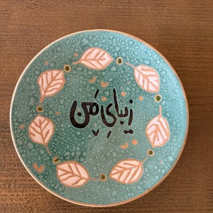 May include: A small, round, turquoise ceramic plate with a textured surface. The plate features a ring of stylized leaves in white and gold around the edge. The center of the plate has black calligraphy.
