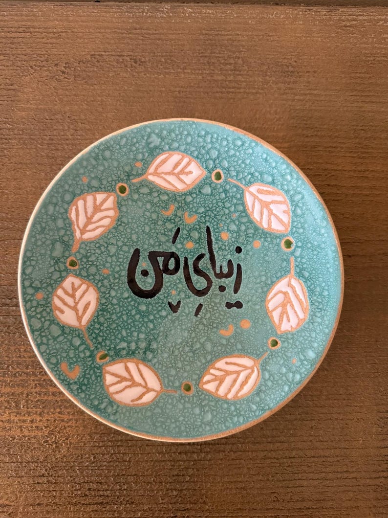 May include: A small, round, turquoise ceramic plate with a textured surface. The plate features a ring of stylized leaves in white and gold around the edge. The center of the plate has black calligraphy.