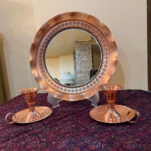 May include: A copper-toned mirror with a decorative floral border is displayed with two matching candle holders. The candle holders have a small handle and a round base. The items are on a patterned purple tablecloth.