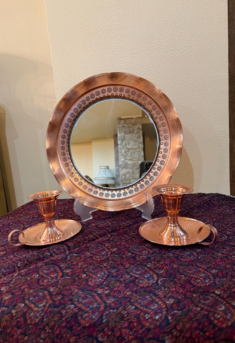 May include: A copper-toned mirror with a decorative floral border is displayed with two matching candle holders. The candle holders have a small handle and a round base. The items are on a patterned purple tablecloth.