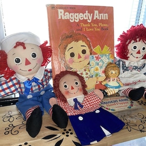 以下が含まれることがあります： 「Raggedy Ann: A Thank You, Please, and I Love You Book」というタイトルの本と、ラグドール人形のコレクション。人形は赤い糸の髪、ペイントされた顔、さまざまな衣装をしています。本はヴィンテージのゴールデンブックです。