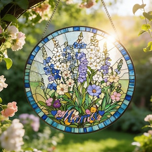 Op de afbeelding: Een rond gebrandschilderd glazen raam met een bloemenontwerp. Het toont blauwe, paarse en witte bloemen met het woord "Mama" in het midden. Het hangt aan een ketting en wordt omgeven door een tuin.