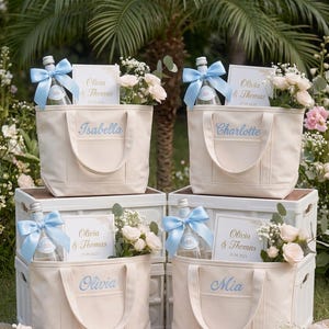 Pode incluir: Quatro sacolas de lona na cor off-white, cada uma personalizada com um nome bordado em azul. Cada sacola contém uma garrafa de vidro com um laço de cetim azul claro, um cartão branco impresso "Olivia & Thomas 25.09.2025" e um pequeno buquê de rosas rosa pálido.